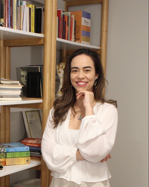 Psicóloga Simone de Siqueira sorrindo ao lado da estante de livros do consultório na Vila Mariana